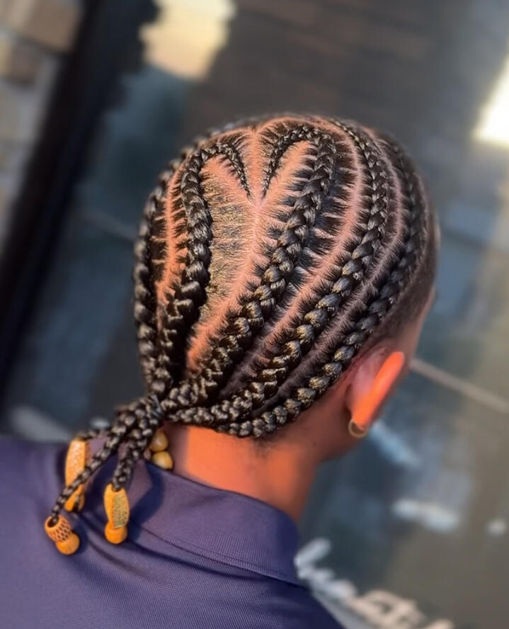Back view of a person with neatly braided cornrows styled into a heart-shaped pattern, tied at the ends with gold accessories.